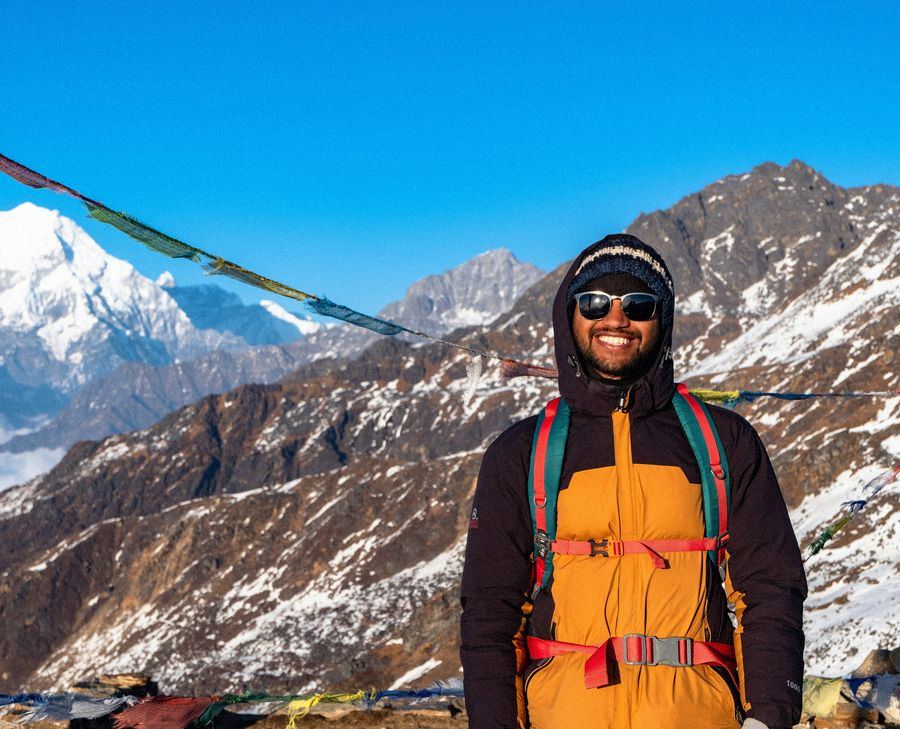A trekker in an orange jacket smiling beneath strings of prayer flags