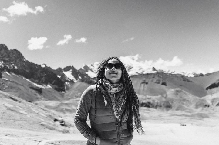 A trekker in sunglasses and a scarf, peaks behind, in black and white